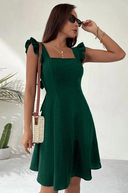 Donna con abito verde elegante, spalline a balze, occhiali da sole, borsa a tracolla intrecciata, sfondo bianco minimalista, pianta decorativa.