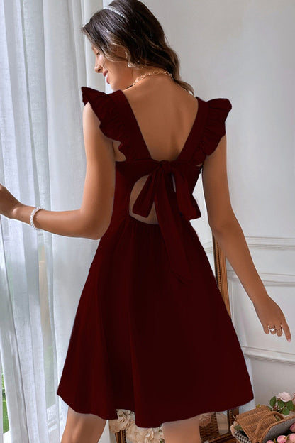 Donna in abito rosso bordeaux con fiocco posteriore, maniche a volant, in piedi vicino a una finestra. Moda elegante, abbigliamento femminile.