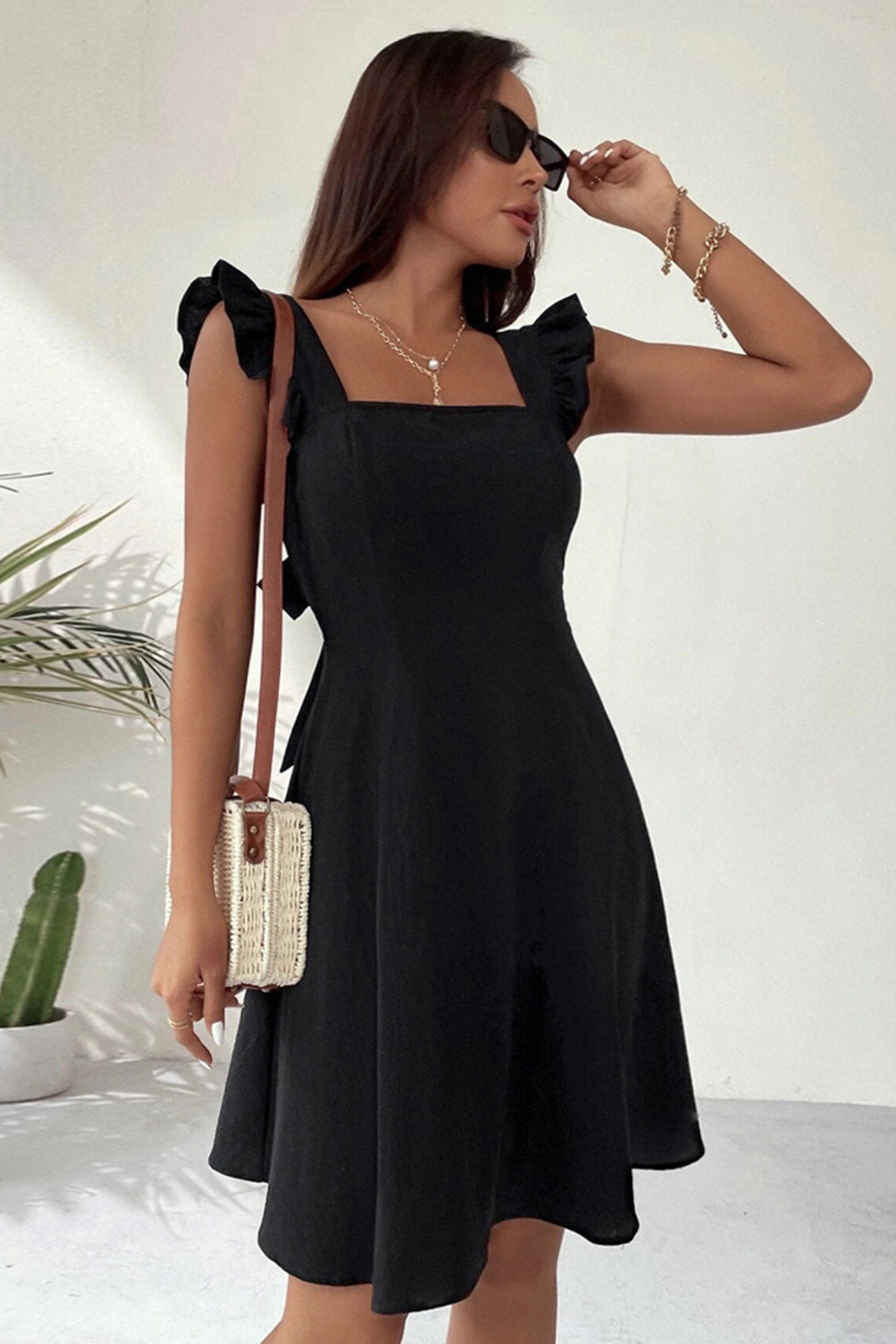 Donna con abito nero estivo, spalline a balze, occhiali da sole, borsa a tracolla in paglia, sfondo bianco minimalista, pianta decorativa.