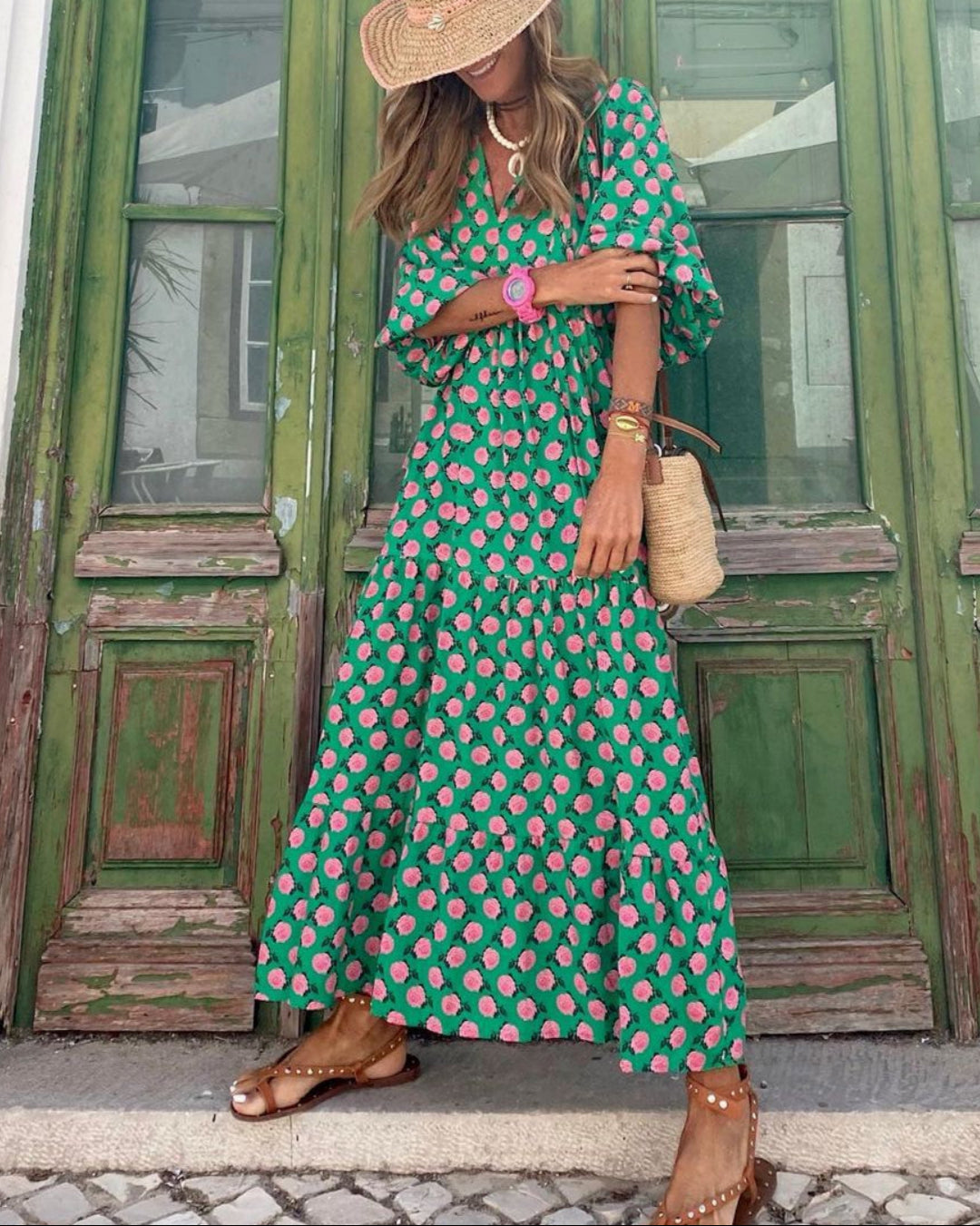 Donna con abito lungo verde a fiori rosa, cappello di paglia, sandali marroni e borsa di paglia, davanti a porte verdi vintage. Moda estiva elegante.