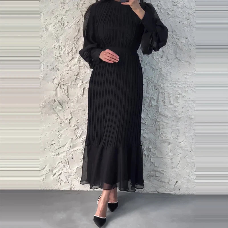 Abito lungo nero elegante con maniche lunghe e gonna plissettata, indossato da una donna. Sfondo muro bianco. Scarpe nere a punta.