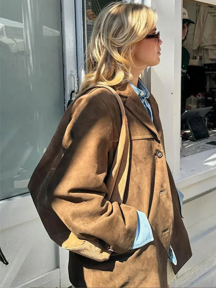 Donna bionda con occhiali da sole indossa giacca di pelle marrone e camicia di jeans, passeggiando all'aperto. Moda autunnale, stile casual chic.