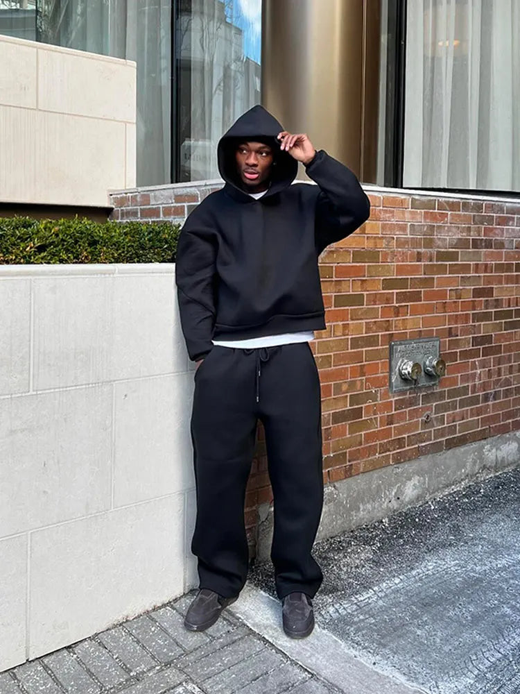 Uomo in abbigliamento casual nero, felpa con cappuccio e pantaloni, posa all'aperto vicino a un edificio in mattoni. Moda urbana, streetwear.