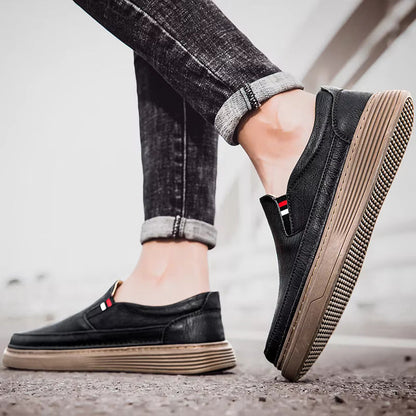 Scarpe slip-on nere da uomo, suola marrone, indossate con jeans scuri arrotolati. Stile casual, moda urbana, comfort quotidiano.