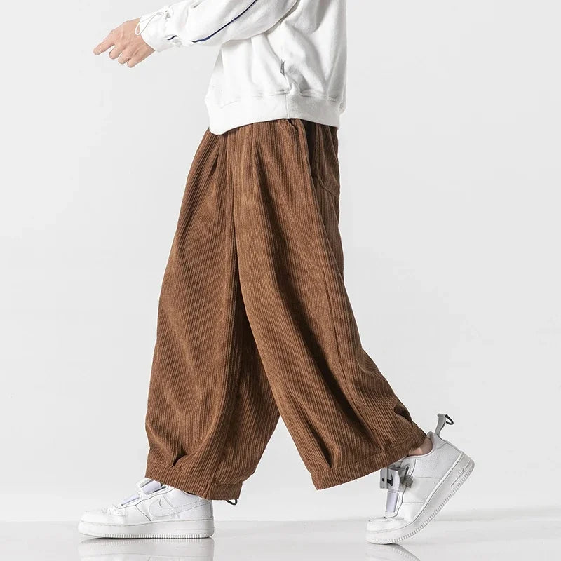 Pantaloni larghi in velluto a coste marroni, indossati con felpa bianca e sneakers bianche. Moda casual uomo, stile urbano contemporaneo.