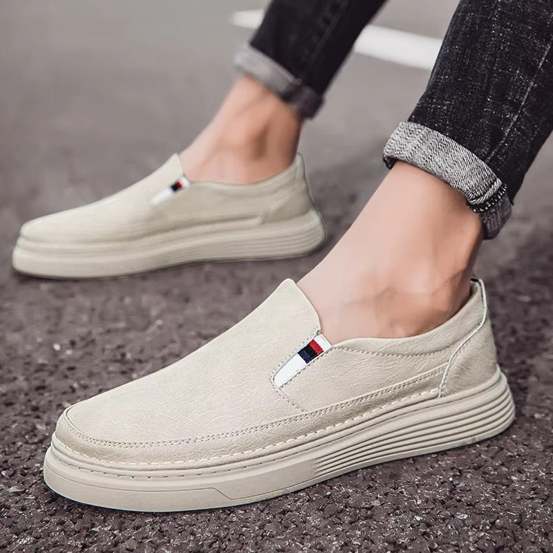 Scarpe slip-on beige da uomo su strada asfaltata, design casual con suola spessa, dettagli tricolore, ideali per moda urbana e comfort quotidiano.