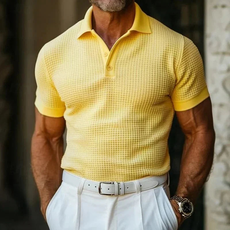 Uomo in polo gialla a trama waffle, pantaloni bianchi eleganti, cintura bianca, orologio da polso. Moda estiva maschile, stile casual chic.