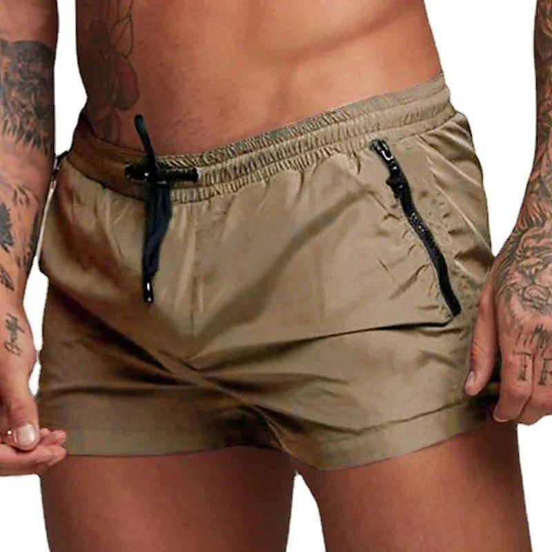 Uomo con tatuaggi indossa pantaloncini da bagno marroni con tasche e cerniere nere, moda estiva, abbigliamento mare maschile.