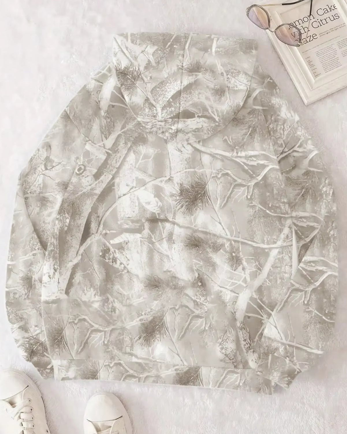 Felpa con cappuccio mimetica grigia, design foresta invernale, su sfondo bianco. Scarpe bianche e occhiali da sole accanto a un libro.