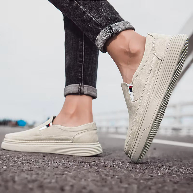 Scarpe slip-on beige da uomo su strada asfaltata, jeans scuri. Calzature casual eleganti, suola spessa, stile urbano moderno.