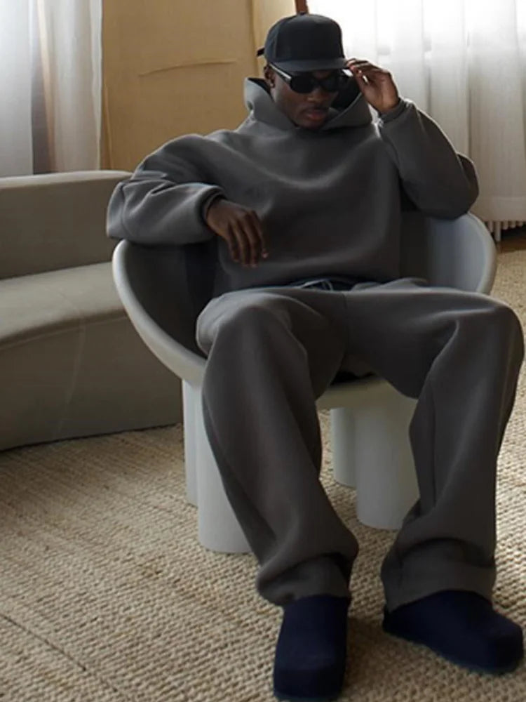 Uomo seduto su sedia moderna, indossa abbigliamento casual grigio, cappello e occhiali da sole. Interno elegante, stile minimalista.