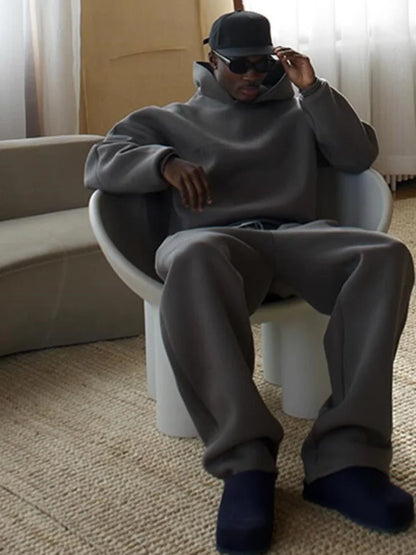 Uomo seduto su sedia moderna, indossa abbigliamento casual grigio, cappello e occhiali da sole. Interno elegante, stile minimalista.
