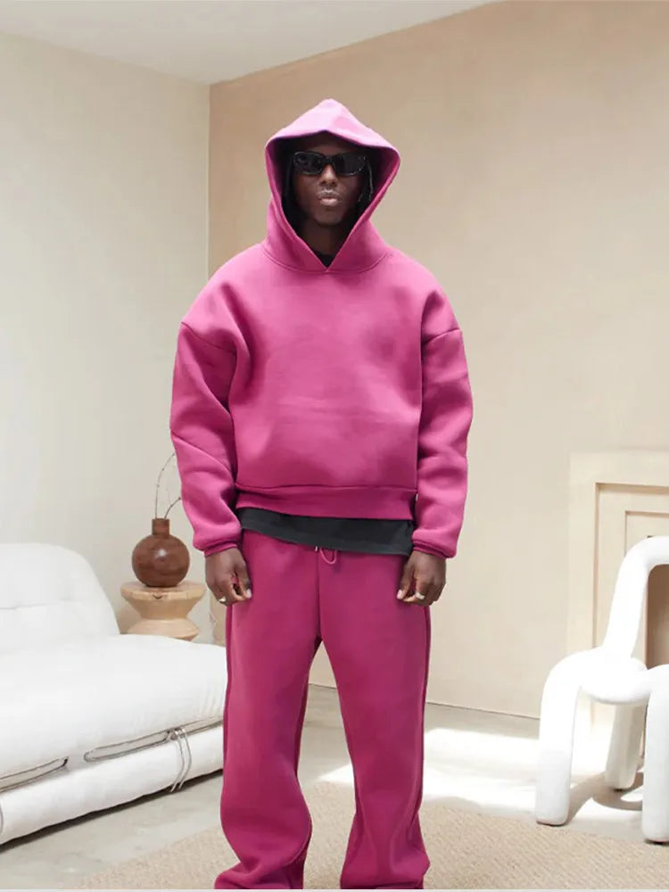 Uomo in tuta sportiva rosa con cappuccio e occhiali da sole, in un salotto moderno. Abbigliamento casual, moda streetwear, stile urbano.