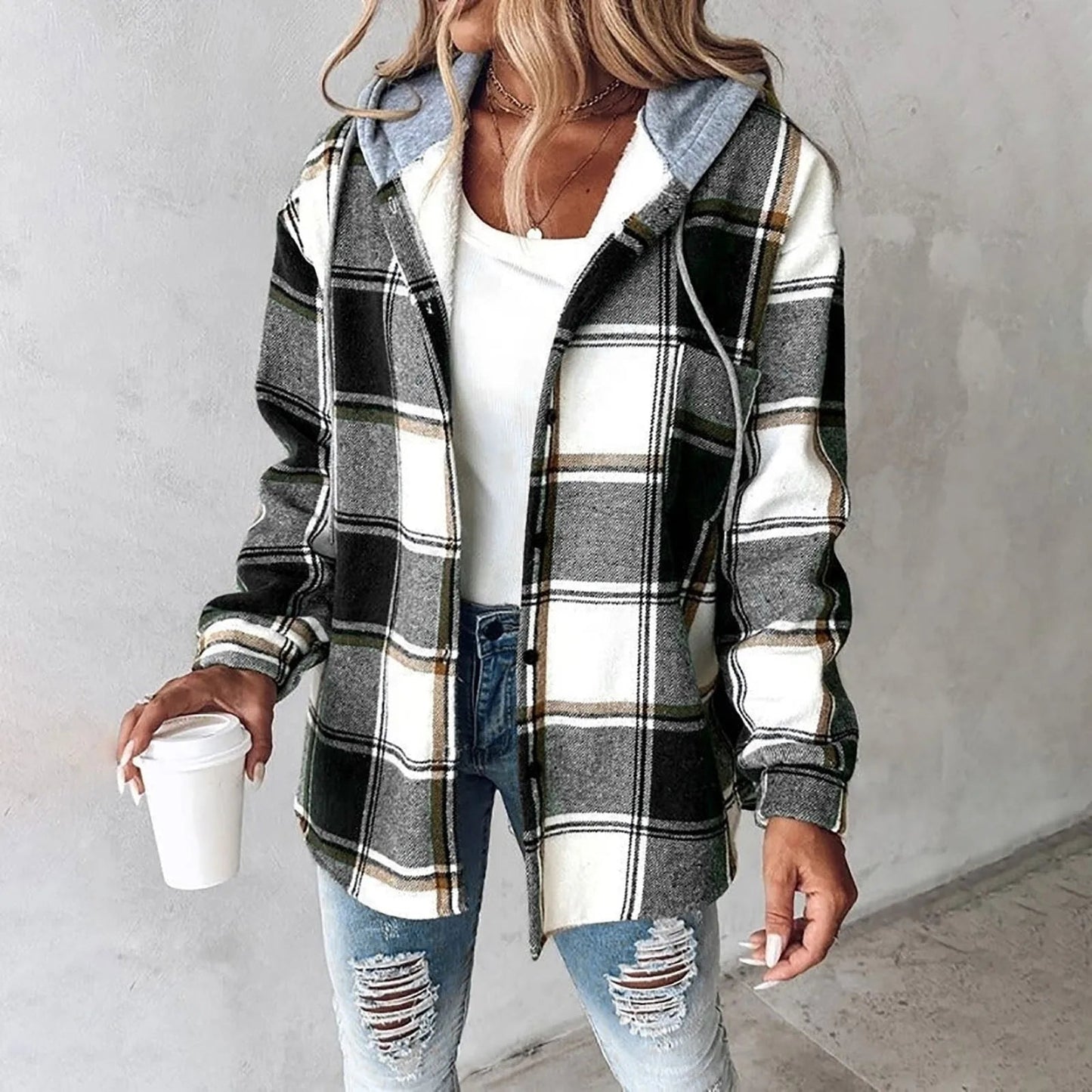 Donna con camicia a quadri bianca e nera, cappuccio grigio, jeans strappati e maglietta bianca, tiene una tazza di caffè. Moda casual autunnale.