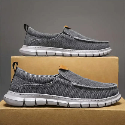Scarpe slip-on grigie da uomo su scatola di cartone, suola bianca, design casual e traspirante, ideali per passeggiate e tempo libero.