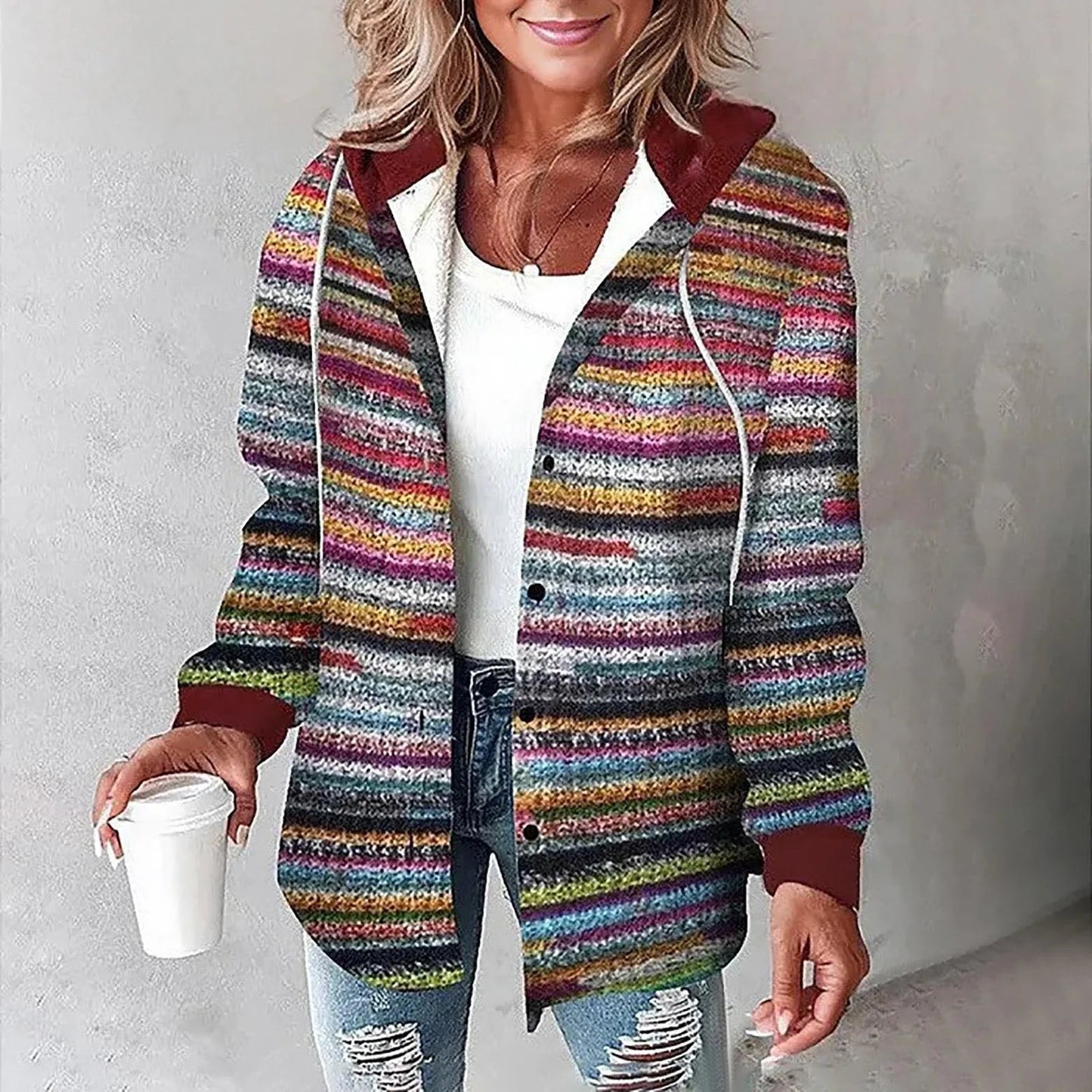 Donna con cardigan a righe multicolore, cappuccio rosso, maglia bianca e jeans strappati, tiene una tazza di caffè. Moda casual autunnale.