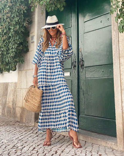 Donna con abito lungo a rombi blu e bianchi, cappello Panama, occhiali da sole e borsa di paglia, davanti a una porta verde. Moda estiva elegante.