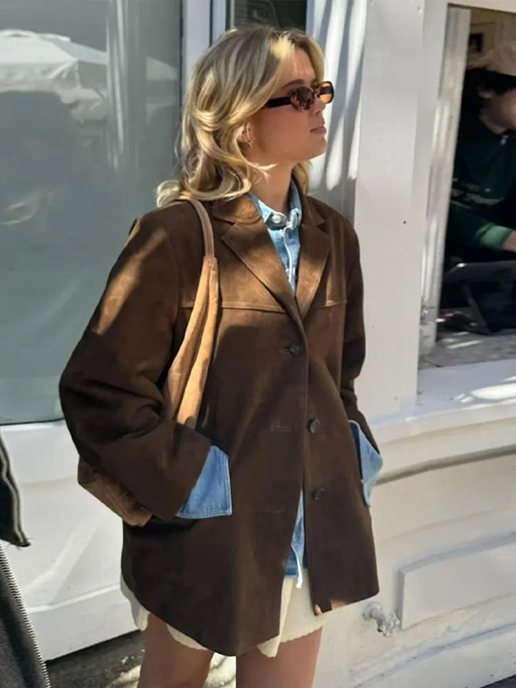 Donna con occhiali da sole indossa giacca marrone su camicia di jeans, borsa a tracolla beige. Moda autunnale, street style, abbigliamento casual.