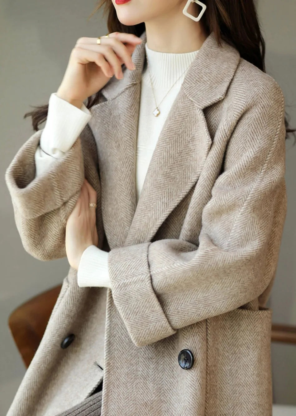 Donna con cappotto beige elegante, maglione bianco a collo alto, orecchini geometrici e anello. Moda autunnale, stile sofisticato e moderno.