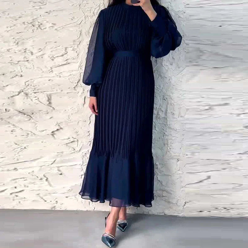 Abito lungo blu scuro con maniche a sbuffo e gonna plissettata, indossato da una donna. Sfondo bianco testurizzato. Moda elegante e sofisticata.