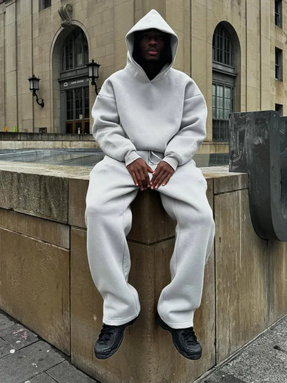 Uomo con felpa e pantaloni grigi seduto su un muretto in città, indossa scarpe nere. Sfondo con edificio storico e lampioni. Moda urbana casual.
