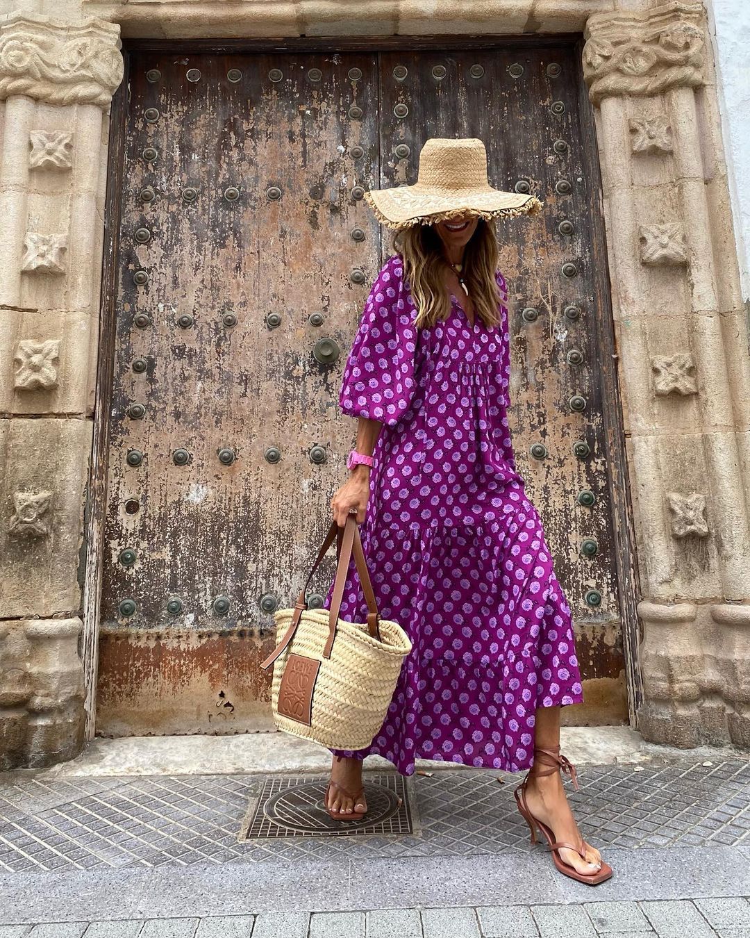 Donna con abito viola a fantasia, cappello di paglia e borsa intrecciata davanti a porta antica. Moda estiva, stile boho-chic, look vacanziero.