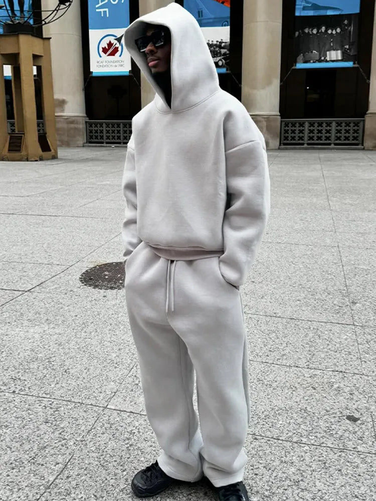 Uomo in tuta grigia con cappuccio e occhiali da sole, in piedi su marciapiede urbano. Moda streetwear, abbigliamento casual, stile moderno.