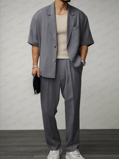 Uomo in abito grigio casual, giacca a maniche corte, pantaloni larghi, maglia beige, scarpe bianche, orologio argento, sfondo a righe.