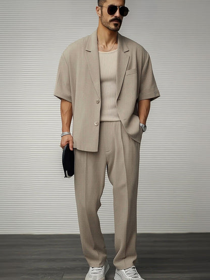 Uomo con completo beige estivo, giacca a maniche corte, pantaloni coordinati, occhiali da sole, orologio, scarpe bianche, sfondo grigio.