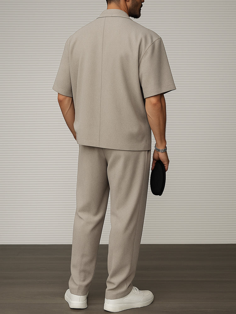 Uomo in abito beige elegante, maniche corte, pantaloni coordinati, scarpe bianche, sfondo a righe grigie. Moda maschile contemporanea.