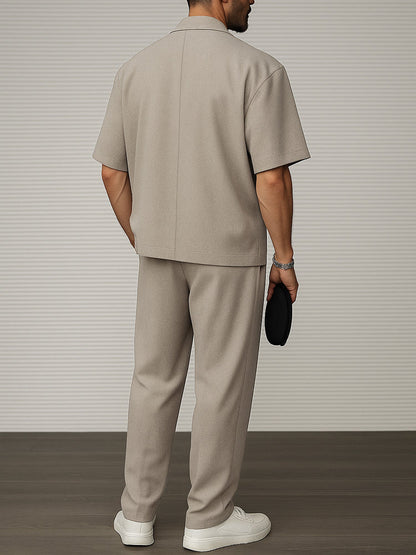 Uomo in abito beige elegante, maniche corte, pantaloni coordinati, scarpe bianche, sfondo a righe grigie. Moda maschile contemporanea.