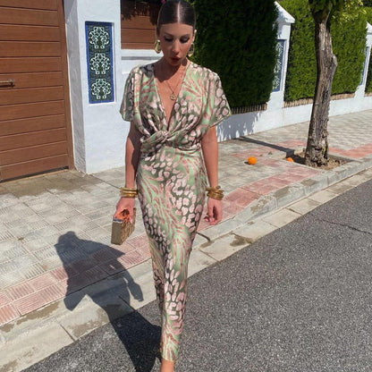 Donna elegante in abito lungo floreale verde e rosa, cammina su marciapiede soleggiato. Accessori dorati, borsa piccola. Moda estiva, stile chic.