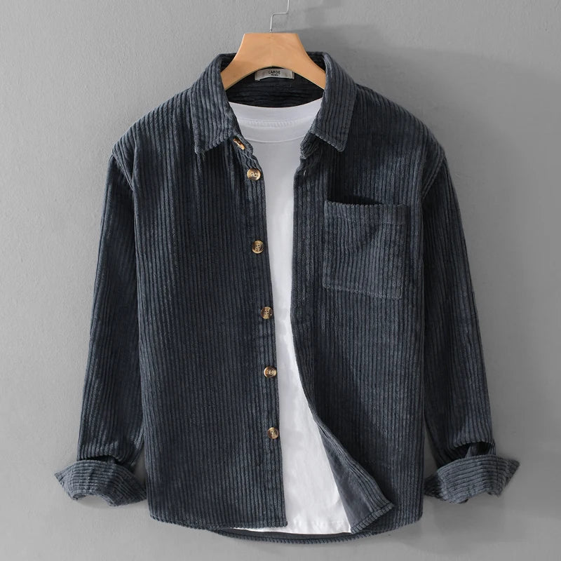 Camicia di velluto a coste grigio scuro su gruccia, con bottoni in legno e taschino, sopra una maglietta bianca. Moda casual uomo.
