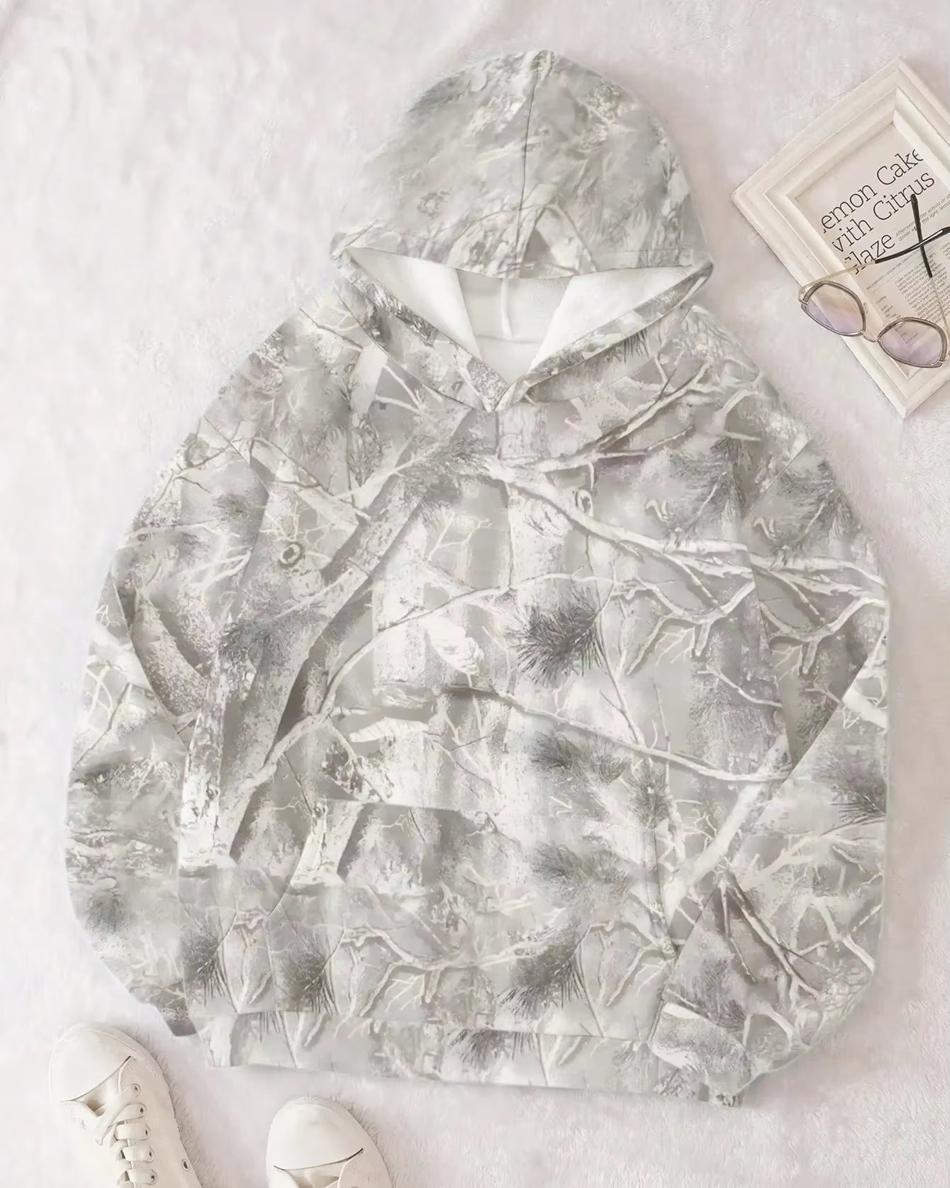 Felpa con cappuccio mimetica bianca e grigia su sfondo chiaro, accanto a occhiali e libro. Design invernale, moda casual, abbigliamento trendy.