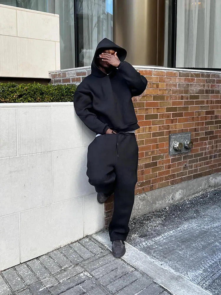 Uomo in abbigliamento sportivo nero con cappuccio, appoggiato a un muro di mattoni, copre il viso con la mano. Urban street style, moda casual.