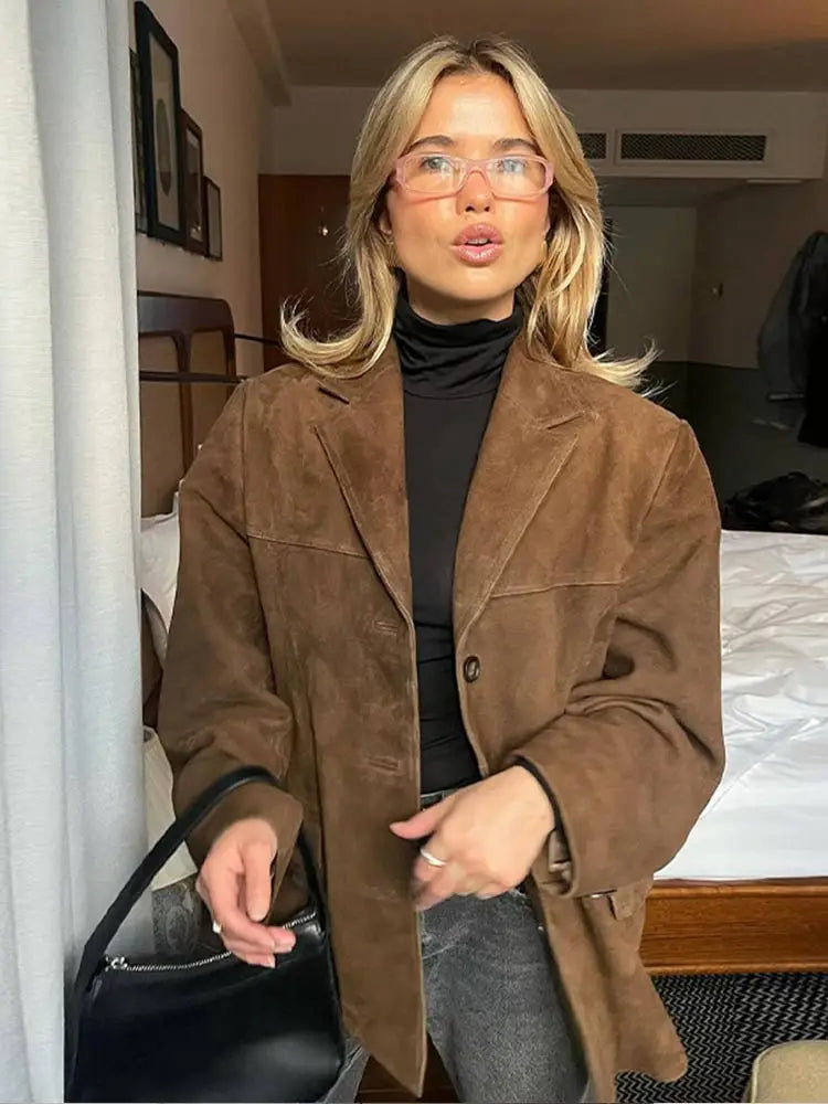 Donna con occhiali e giacca marrone in camera d'albergo, moda autunnale, look casual chic, borsa nera, capelli biondi, stile moderno.