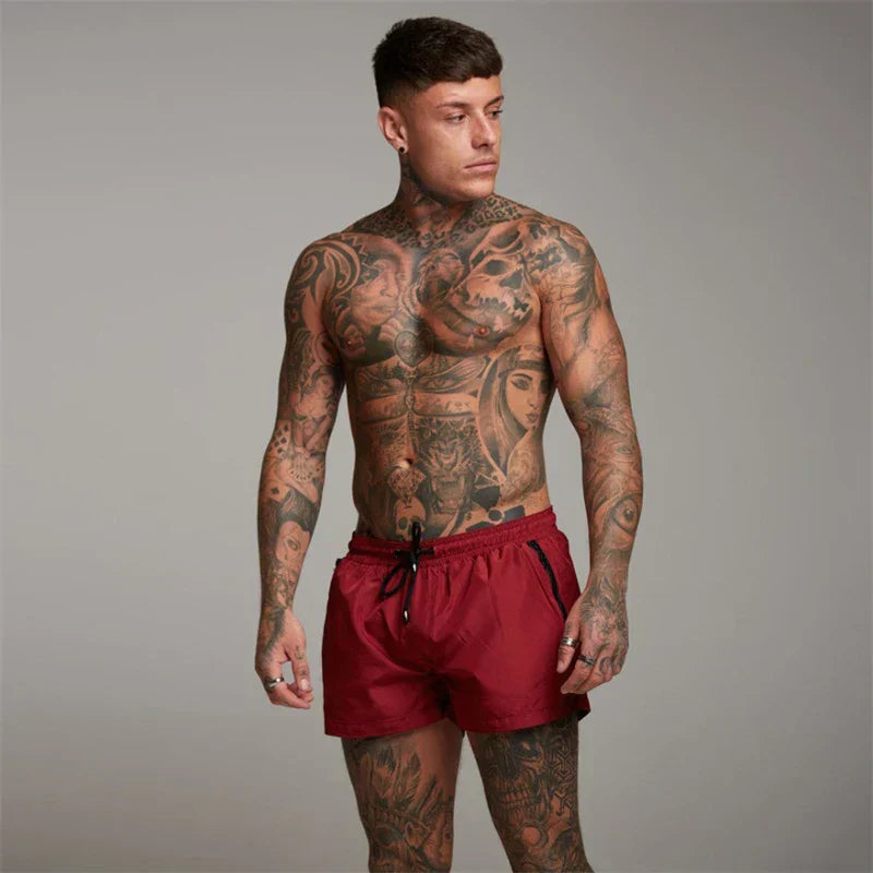 Uomo muscoloso con tatuaggi complessi su tutto il corpo, indossa pantaloncini rossi. Sfondo neutro. Fitness, tatuaggi artistici, moda maschile.