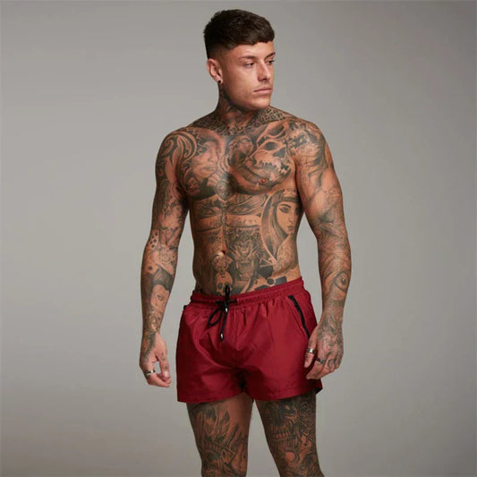 Uomo muscoloso con tatuaggi complessi su tutto il corpo, indossa pantaloncini rossi. Sfondo neutro. Fitness, tatuaggi artistici, moda maschile.