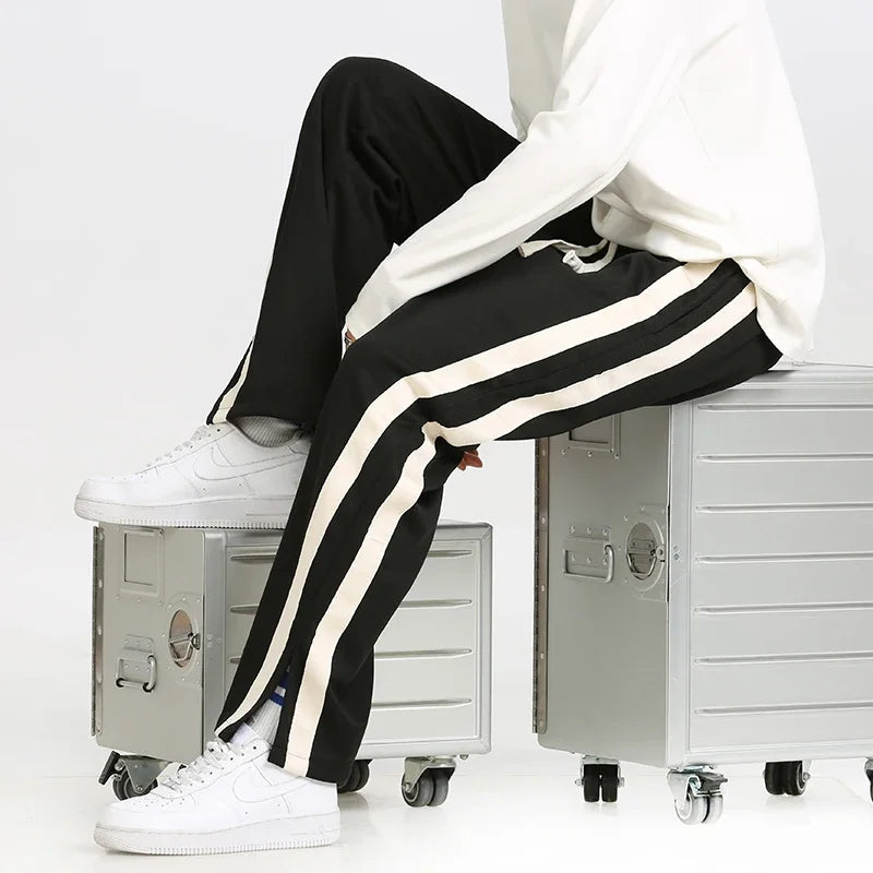 Pantaloni sportivi neri con strisce bianche, indossati con sneakers bianche, seduto su valigie metalliche. Moda casual uomo, stile urbano.