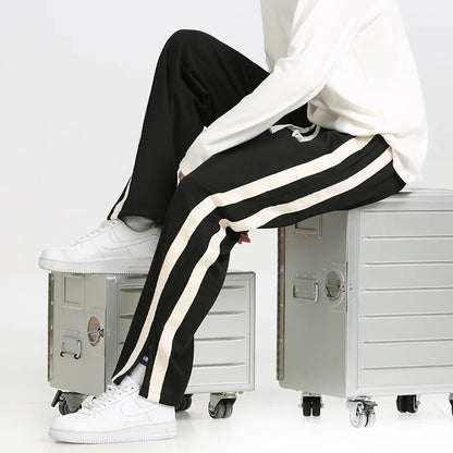 Pantaloni sportivi neri con strisce bianche, indossati con sneakers bianche, seduto su valigie metalliche. Moda casual uomo, stile urbano.