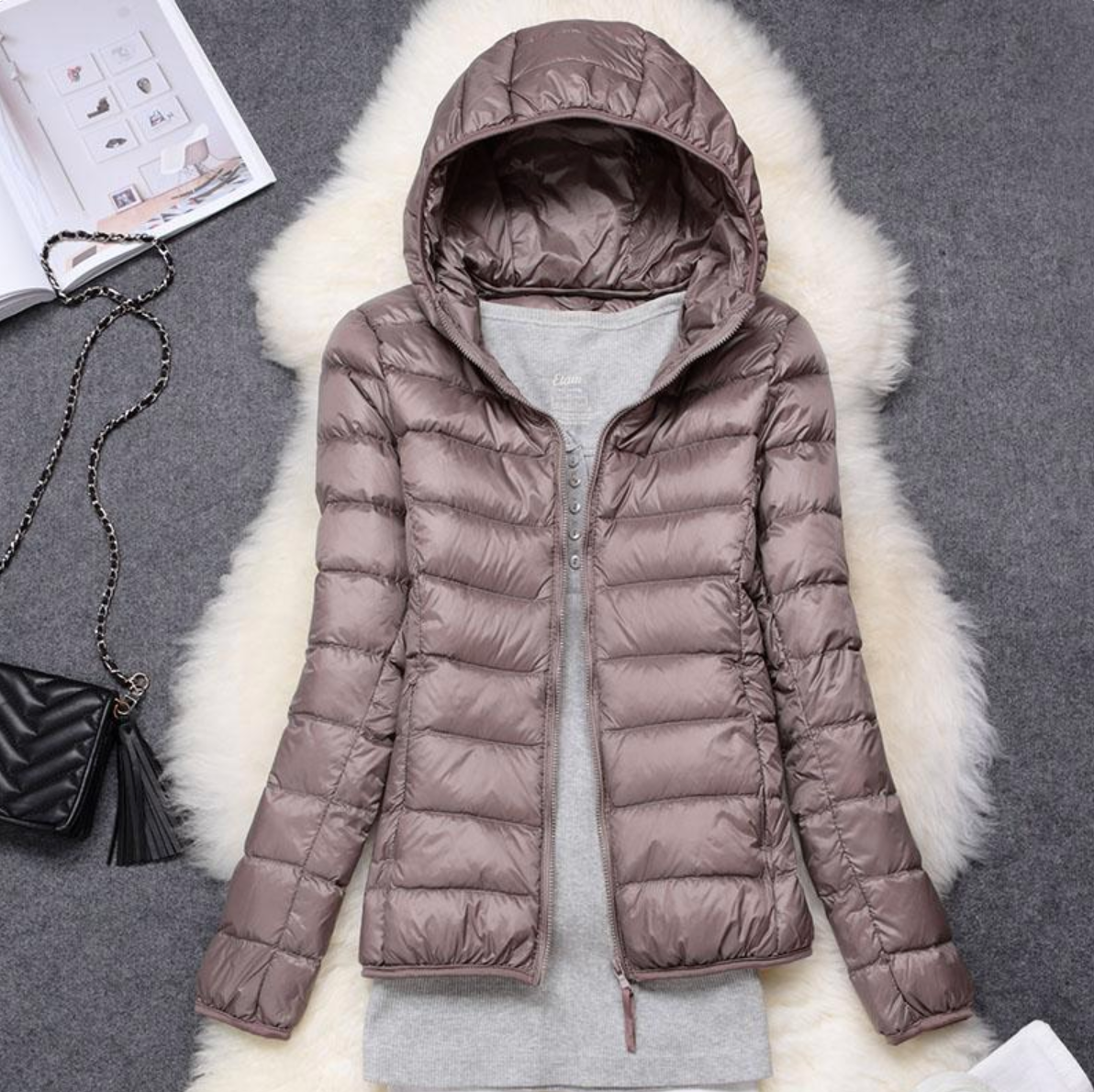 Giacca invernale imbottita marrone con cappuccio, su sfondo grigio, accanto a borsa nera e rivista. Moda donna, abbigliamento caldo, stile casual.
