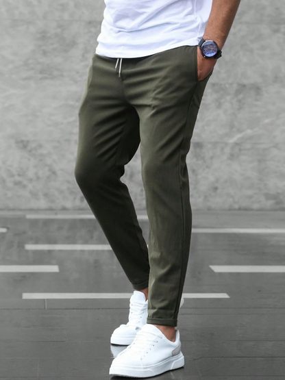 Pantaloni jogger verde oliva da uomo, abbinati a scarpe da ginnastica bianche e maglietta bianca, moda casual urbana, sfondo grigio.