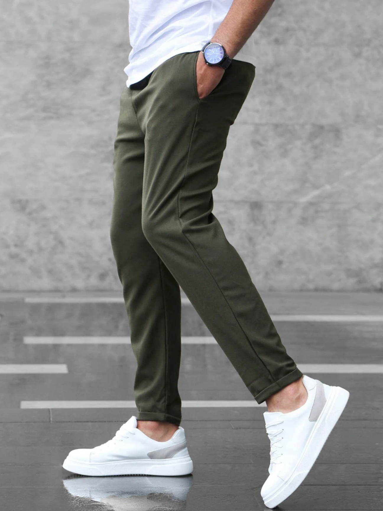 Uomo con pantaloni chino verde oliva, scarpe da ginnastica bianche e orologio blu, cammina su pavimento grigio. Moda casual maschile.