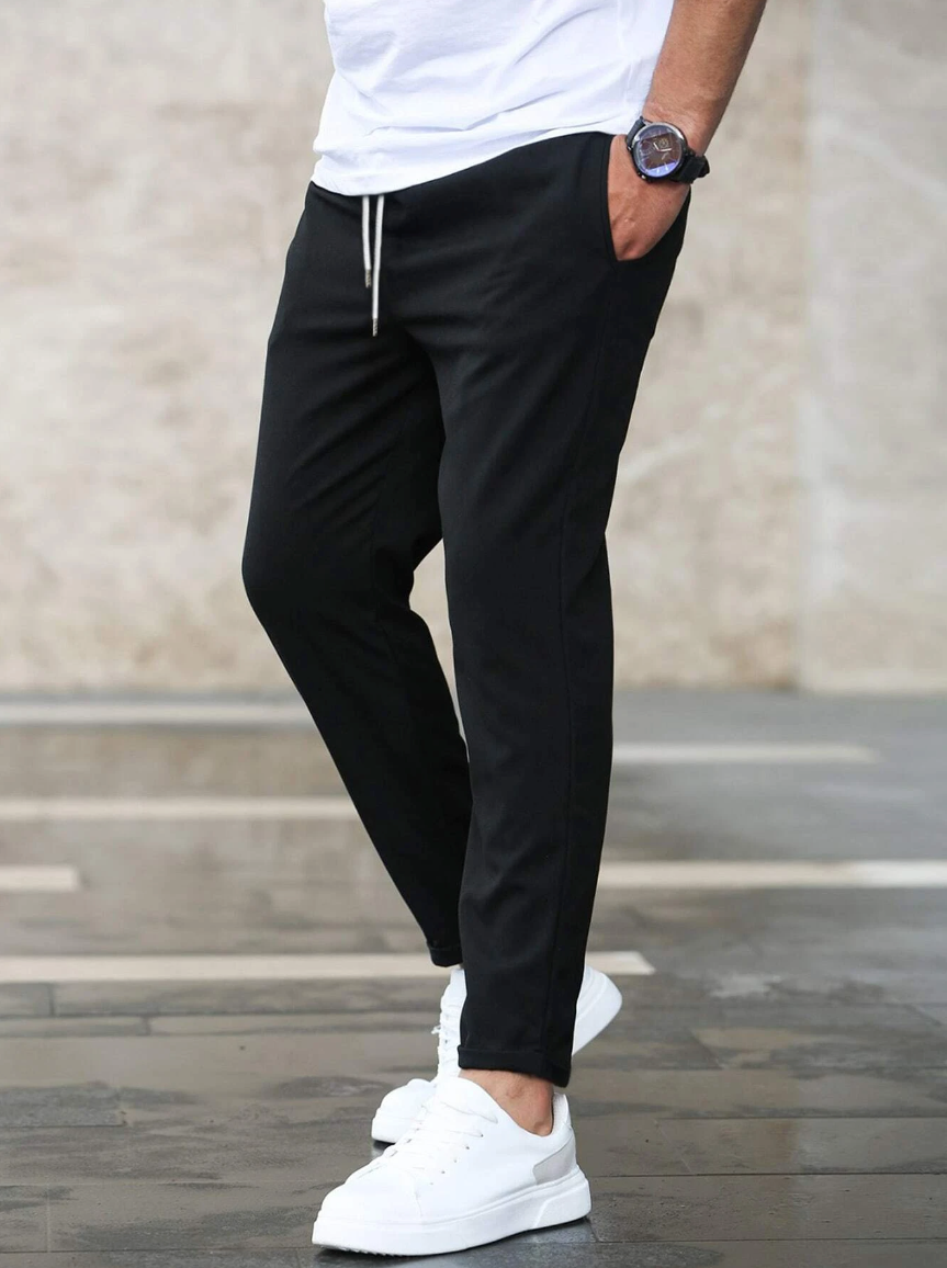 Uomo con pantaloni neri casual e scarpe da ginnastica bianche, orologio al polso, su sfondo urbano. Moda maschile, abbigliamento casual.