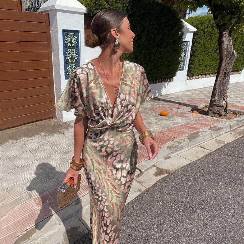 Donna elegante in abito lungo floreale verde e rosa, accessori dorati, cammina su marciapiede soleggiato. Moda estiva, stile chic, look sofisticato.