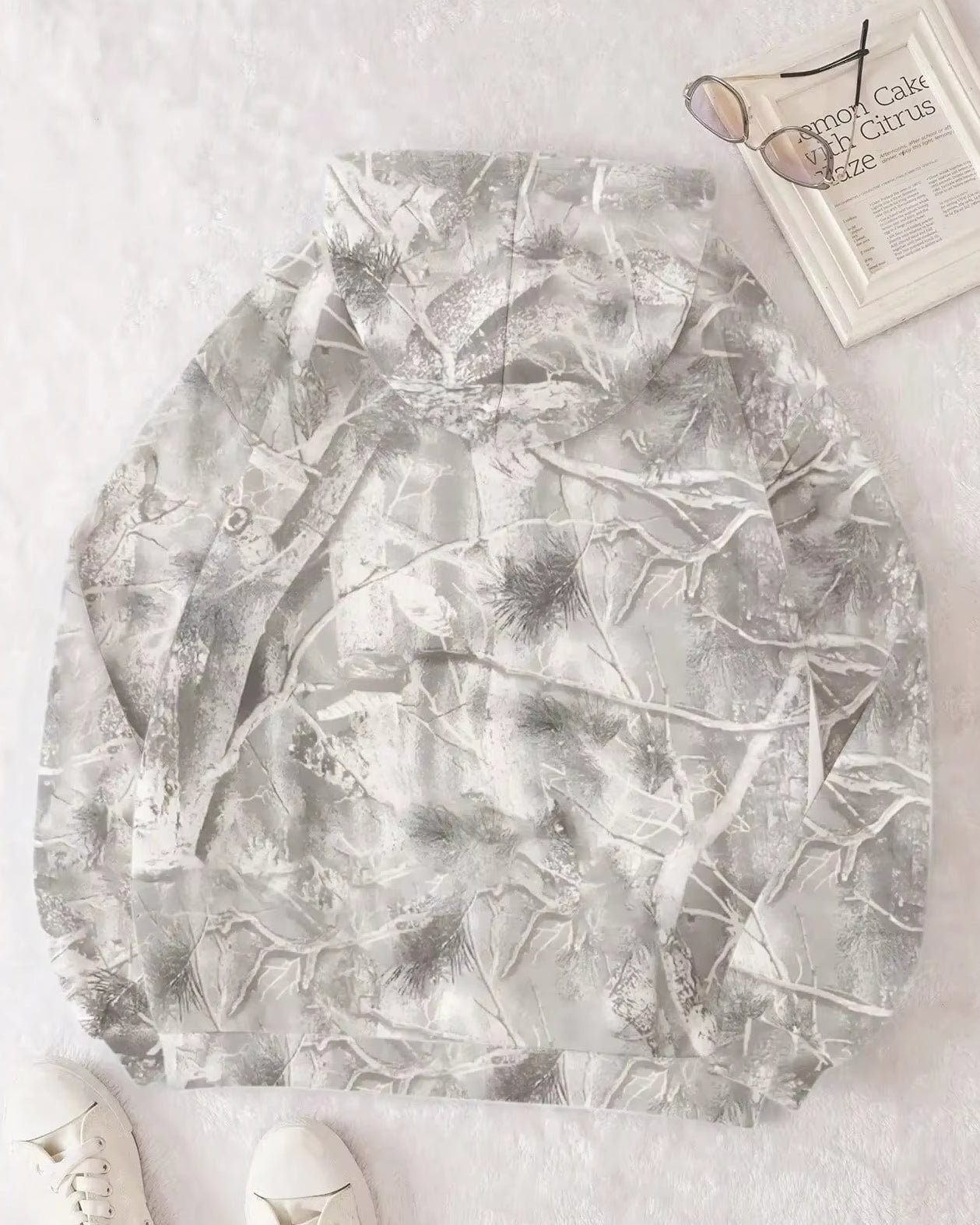 Felpa con cappuccio mimetica grigia, motivo foresta invernale, su sfondo bianco. Include occhiali da sole, rivista e scarpe bianche.