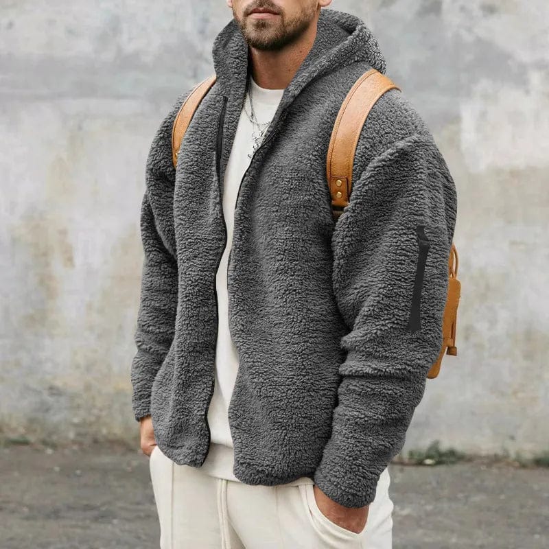 Uomo con giacca in pile grigio, cappuccio e zaino marrone, sfondo urbano. Moda invernale casual, abbigliamento caldo e confortevole.