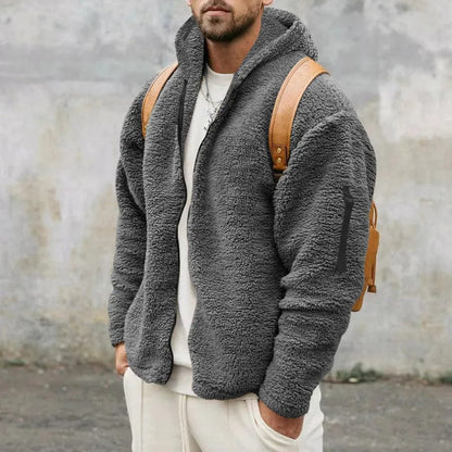 Uomo con giacca in pile grigio, cappuccio e zaino marrone, sfondo urbano. Moda invernale casual, abbigliamento caldo e confortevole.