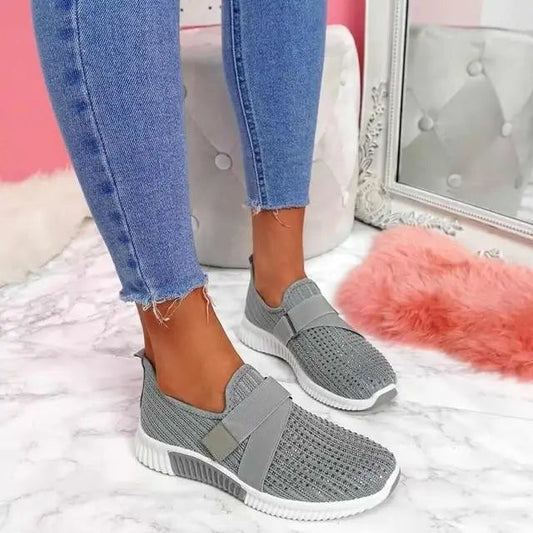 Scarpe da ginnastica grigie in maglia su pavimento in marmo, indossate con jeans aderenti. Design moderno e comodo, perfetto per look casual.