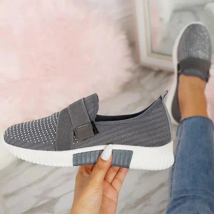 Scarpa sportiva grigia in maglia traspirante con suola bianca, design slip-on, tenuta in mano su sfondo di pavimento marmo e tappeto rosa.